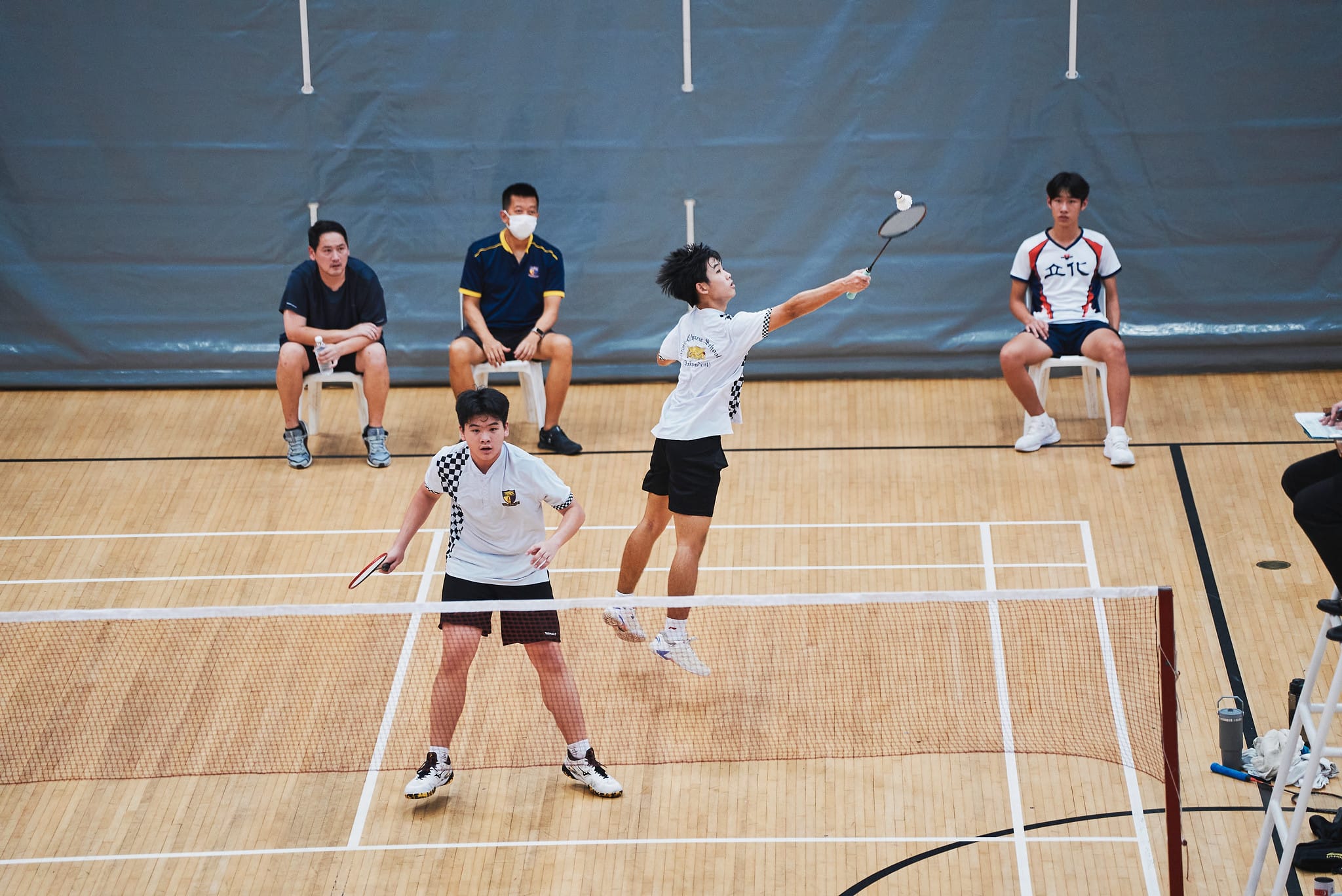A doubles badminton match in progress, with two players on one side of the net in active playing stances on a wooden court.